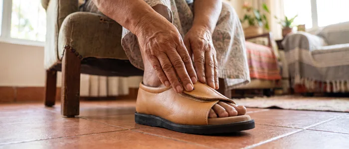 An older woman fastening the wide velcro strap of her tan AJL Majestic open-toe diabetic slip-on shoe at home